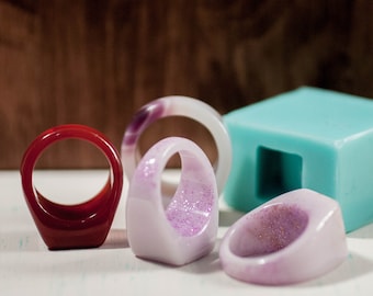 May include: A selection of resin rings in different colours and styles. The rings include a deep red, a light purple with glitter, and a translucent ring with purple marbling. A light blue square object is also present.