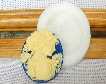 May include: An oval-shaped cameo with a profile of a woman in cream against a blue background. A white silicone mold is next to the cameo. The cameo has a detailed design of a woman's profile with curly hair and floral accents.