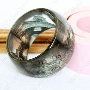 Könnte beinhalten: Ein breites, transparentes Armreif-Armband mit einem Graustufenbild des Eiffelturms und der Kathedrale Notre-Dame. Das Armband hat eine dunkle, rauchige Farbe und eine glatte, abgerundete Form. Das Armband hat einen Durchmesser von etwa 6,35 cm.