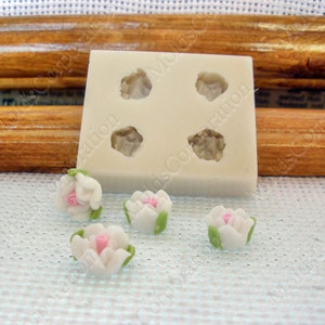 May include: A white silicone mold with four flower-shaped cavities, alongside four miniature white and pink flower decorations with green leaves. The flowers are likely for crafting or cake decorating.