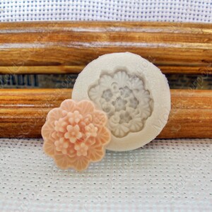 Puede incluir: Un jabón en forma de flor, de color melocotón claro, junto a un molde de silicona blanco. El molde tiene un diseño floral. El jabón es una réplica detallada del patrón del molde. El fondo incluye un marco de madera y tela blanca.