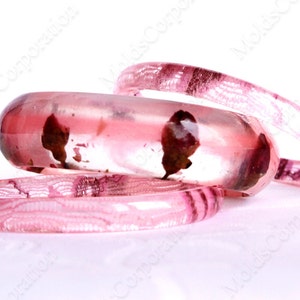 Puede incluir: Tres pulseras de resina transparente en tonos rosa y granate. La más grande tiene forma redondeada, las otras son más finas. La resina contiene inclusiones granates, creando un diseño único. Adecuado para una circunferencia de muñeca de unos 18 cm.