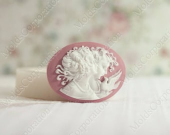May include: A pink oval cameo mold featuring a white profile of a woman with curly hair and a dove. The cameo is set against a soft, blurred background. The words "Molds Corporation" are visible in the background.