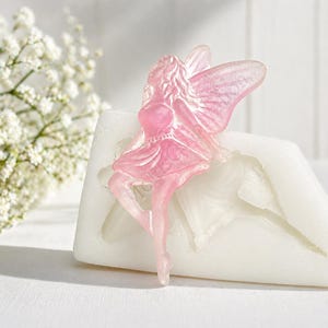 May include: A pink fairy figurine with translucent wings and a flowing dress. The fairy is posed gracefully, and is set against a white background. The fairy is a decorative item.