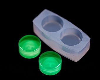 May include: A clear, light blue silicone mold with two circular cavities, next to two glowing green resin circles. The resin circles have a translucent appearance, and the mold is rectangular with rounded edges. The background is black.