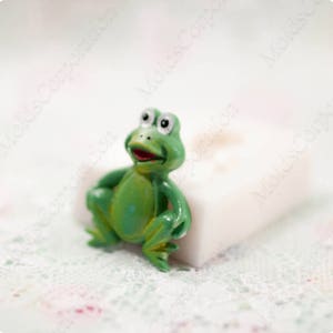 Può includere: Una piccola statuetta a forma di rana verde con un'espressione sorridente e la bocca rossa. La rana ha grandi occhi bianchi e posa con le braccia lungo i fianchi. La statuetta è su una superficie bianca.