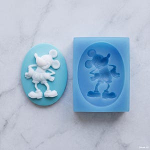 May include: A light blue oval-shaped silicone mold featuring a recessed design of a cartoon mouse character with large round ears, wearing shorts and shoes. Beside the mold is a white cast of the same character, showing the raised relief detail.
