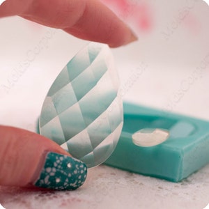 May include: A clear, faceted, teardrop-shaped resin piece with a light blue and white diamond pattern, held between fingers. A small, similar piece and a teal mold are in the background. The image is well-lit, highlighting the details.