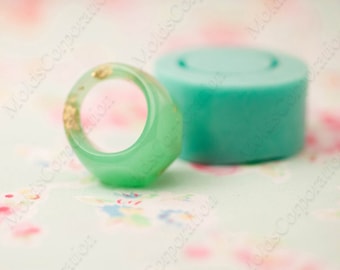 May include: A translucent green resin ring with gold leaf inclusions. Beside it is a cylindrical teal mould. The ring has a rounded shape and a smooth surface. The background features a floral pattern.