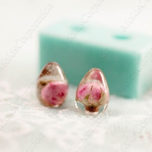 May include: Two clear resin teardrop-shaped earrings, each containing a pink rose bud. The earrings sit on a white lace surface, with a light blue rectangular object in the background. The earrings are a unique floral accessory.