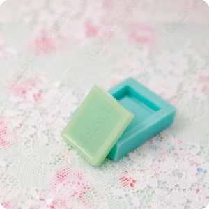 Puede incluir: Una pastilla de jabón rectangular verde claro junto a un molde rectangular turquesa. La pastilla de jabón tiene bordes redondeados y el texto "Molds Corporation" es visible. El molde tiene forma cuadrada con el interior rebajado.