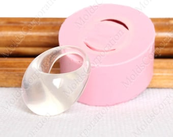 May include: A clear, oval ring sits beside a pink, circular mould. The ring is transparent, allowing light to pass through. The mould has a curved indentation, mirroring the ring's shape. The background is a light colour.