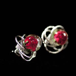 Könnte beinhalten: Ein Paar silberfarbene Ohrstecker. Jeder Ohrring hat eine klare, kugelförmige Harzkuppel, die eine leuchtend rote Rose umschließt. Die Ohrringe haben ein verdrehtes, verwobenes Design um die Harzkuppel, vor einem schwarzen Hintergrund.