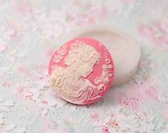 May include: A pink cameo mold with a white relief of a woman's profile, surrounded by floral details. The mold is round and sits next to a white, oval-shaped mold. The background is a floral lace pattern.