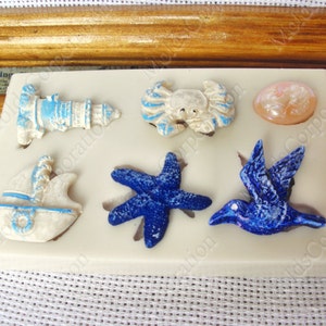 May include: A silicone mold featuring nautical-themed shapes. Includes a lighthouse, crab, seashell, sailboat, starfish, and a bird. The shapes are white with blue accents and are arranged on a beige surface.