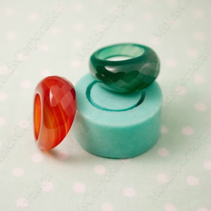 May include: Two faceted gemstone rings, one red and one green, sit next to a teal-colored mold. The red ring has orange and yellow striations. The green ring is a deep emerald color. The rings are on a light green polka dot background.