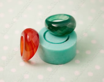 May include: Two faceted gemstone rings, one red and one green, sit next to a teal-coloured mould. The red ring has orange and yellow striations. The green ring is a deep emerald colour. The rings are on a light green polka dot background.