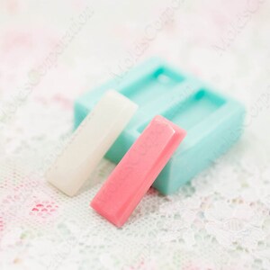 Op de afbeelding: Een lichtblauwe siliconen mal met twee rechthoekige holtes, naast twee rechthoekige staven, één witte en één roze. De staven lijken zeep of hars te zijn. De achtergrond is een bloemen kanten patroon.