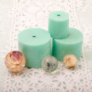Puede incluir: Una colección de moldes cilíndricos de color turquesa de diferentes alturas, junto con tres esferas decorativas. Las esferas presentan flores secas y elementos naturales encapsulados en resina transparente, sobre un fondo de encaje blanco.