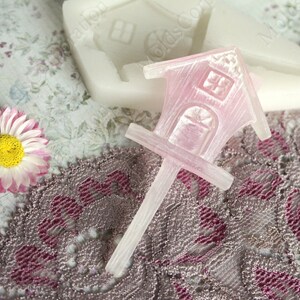 Può includere: Uno stampo a forma di casetta per uccelli rosa traslucido con un lungo stelo, probabilmente per l'artigianato. La casetta per uccelli ha una porta e una finestra. Uno stampo bianco e un fiore rosa sono sullo sfondo, su un tessuto fantasia.