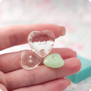 Op de afbeelding: Drie hartvormige knopen in verschillende maten en kleuren. Twee zijn helder met facetgeslepen oppervlakken en één is zachtgroen. Elke knoop heeft twee gaten om te naaien. De knopen worden in een hand gehouden.