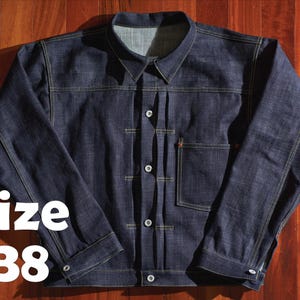 Könnte beinhalten: Dunkelblaue Jeansjacke mit Knopfleiste, einer Brusttasche und einem Kragen. Die Jacke hat gelbe Nähte und den Text "Size 38" in Weiß. Die Jacke wird auf einer Holzoberfläche präsentiert.