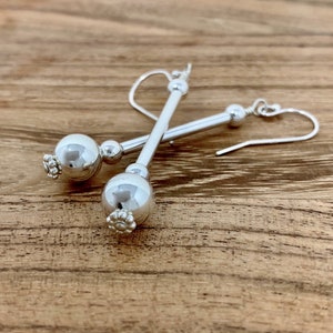 May include: A pair of silver dangle earrings with a round bead and a smaller bead at the top. The earrings are on a wooden surface.