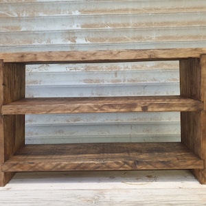 May include: A three-tiered wooden shelf unit with a rustic appearance. The shelves and supports are made from thick, dark-stained wood, displaying the natural grain. The unit has a simple, robust design, ideal for storage or display purposes.