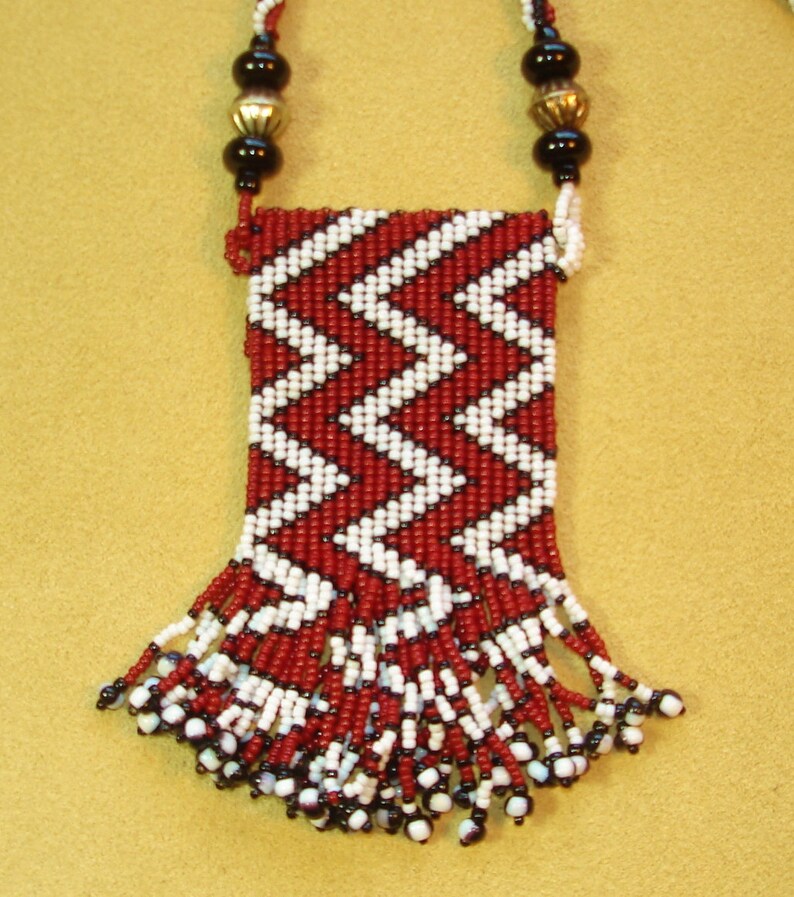 Puede incluir: Un collar de cuentas rojas y blancas con flecos y una peque&ntilde;a bolsa. La bolsa est&aacute; decorada con un patr&oacute;n de chevron.