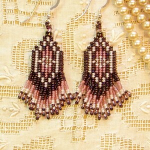 Peut inclure: Paire de boucles d'oreilles perl&eacute;es faites &agrave; la main avec un motif floral. Les boucles d'oreilles pr&eacute;sentent une combinaison de perles marron fonc&eacute;, roses et blanches, avec des crochets d'oreilles argent&eacute;s. Elles sont pr&eacute;sent&eacute;es sur un tissu en dentelle cr&egrave;me.