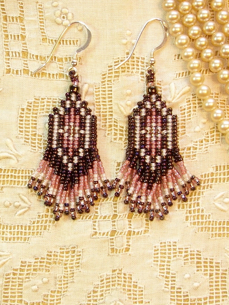 Peut inclure: Paire de boucles d'oreilles perl&eacute;es faites &agrave; la main avec un motif floral. Les boucles d'oreilles pr&eacute;sentent une combinaison de perles marron fonc&eacute;, roses et blanches, avec des crochets d'oreilles argent&eacute;s. Elles sont pr&eacute;sent&eacute;es sur un tissu en dentelle cr&egrave;me.