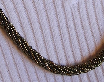 May include: A close-up of a gold-toned chain necklace made of small beads. The necklace is lying on a white textured surface.