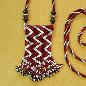 Puede incluir: Un collar de cuentas rojas y blancas con una peque&ntilde;a bolsa adjunta. La bolsa est&aacute; decorada con un patr&oacute;n de chevron y tiene flecos.