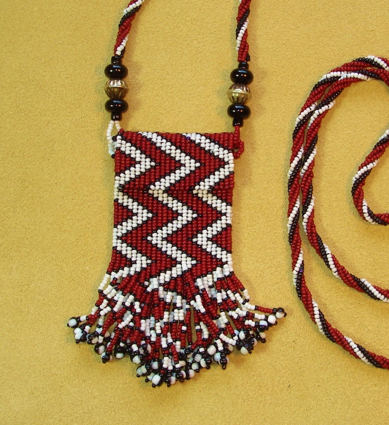 Puede incluir: Un collar de cuentas rojas y blancas con una peque&ntilde;a bolsa adjunta. La bolsa est&aacute; decorada con un patr&oacute;n de chevron y tiene flecos.