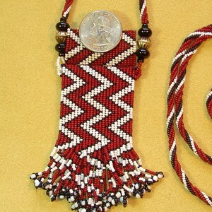 Puede incluir: Un collar de cuentas rojas y blancas con una peque&ntilde;a bolsa y flecos. La bolsa est&aacute; decorada con un patr&oacute;n de chevron. El collar mide aproximadamente 45 cm de largo.