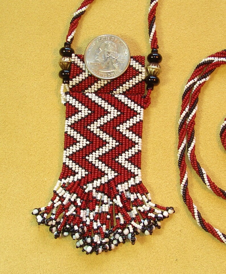Puede incluir: Un collar de cuentas rojas y blancas con una peque&ntilde;a bolsa y flecos. La bolsa est&aacute; decorada con un patr&oacute;n de chevron. El collar mide aproximadamente 45 cm de largo.