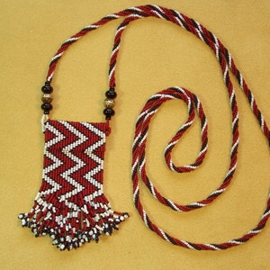 Puede incluir: Un collar de cuentas rojas, blancas y negras con una bolsa de cuentas a juego. La bolsa tiene flecos y est&aacute; unida al collar.