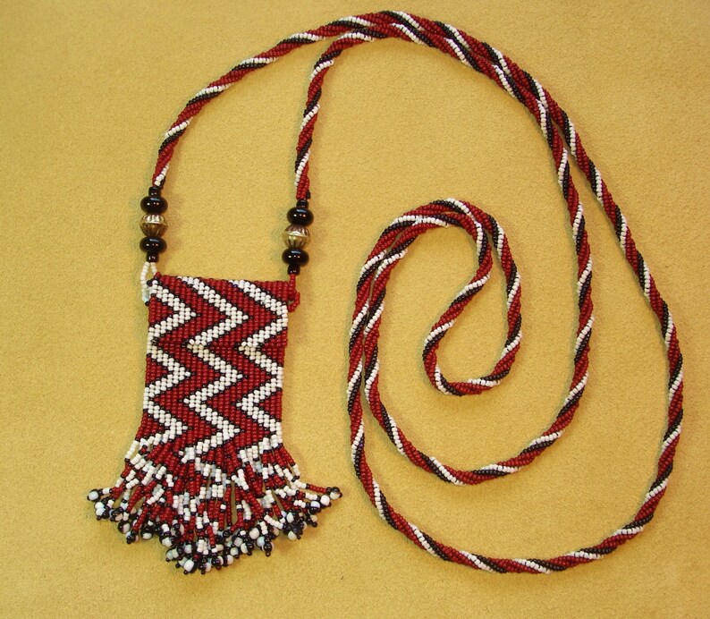 Puede incluir: Un collar de cuentas rojas, blancas y negras con una bolsa de cuentas a juego. La bolsa tiene flecos y est&aacute; unida al collar.