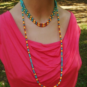 Puede incluir: Un collar largo de cuentas turquesa, naranja y amarillo con un diseño en capas. El collar lo lleva una persona que lleva una camisa rosa.