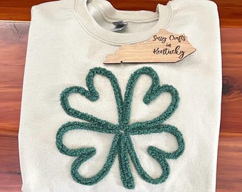 May include: A light green sweatshirt with a four-leaf clover design. The clover is made of fluffy, textured fabric. The sweatshirt has a crew neck and is made of a soft, comfortable material. The sweatshirt also features a wooden cutout of the state of Kentucky with the text "Sassy Crafts in Kentucky".