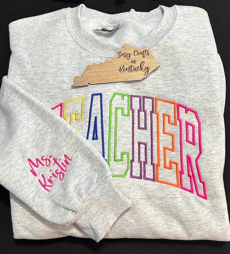 May include: A gray sweatshirt with the word "TEACHER" embroidered in rainbow colors. The word is outlined in black. The sweatshirt has a pink embroidered name on the left sleeve, "Ms. Kristin". A wooden sign with the text "Sassy Crafts in Kentucky" is in the background.