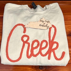 May include: A cream-coloured crewneck sweatshirt featuring the word Creek in large, textured, orange chenille-style script lettering across the front. A wooden cutout shaped like the state of Kentucky with the text Sassy Crafts in Kentucky is placed.