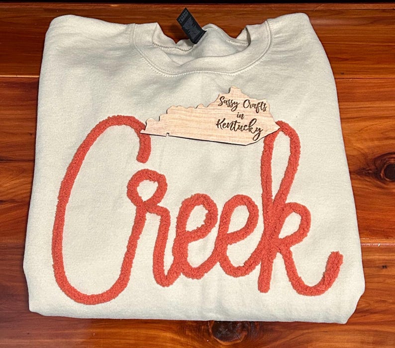 May include: A cream-coloured crewneck sweatshirt featuring the word Creek in large, textured, orange chenille-style script lettering across the front. A wooden cutout shaped like the state of Kentucky with the text Sassy Crafts in Kentucky is placed.