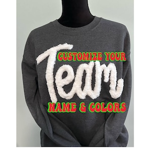 May include: A dark gray sweatshirt with the words "Customize Your Team Name & Colors" in white, green, and red. The word "Team" is in a fluffy, textured font. The sweatshirt is displayed on a mannequin.