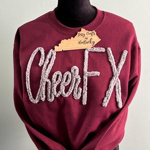 May include: A maroon crewneck sweatshirt featuring the text CheerFX in raised, textured, light grey chenille-style lettering. A wooden cutout shaped like the state of Kentucky with the text Sassy Crafts in Kentucky is placed at the neckline.