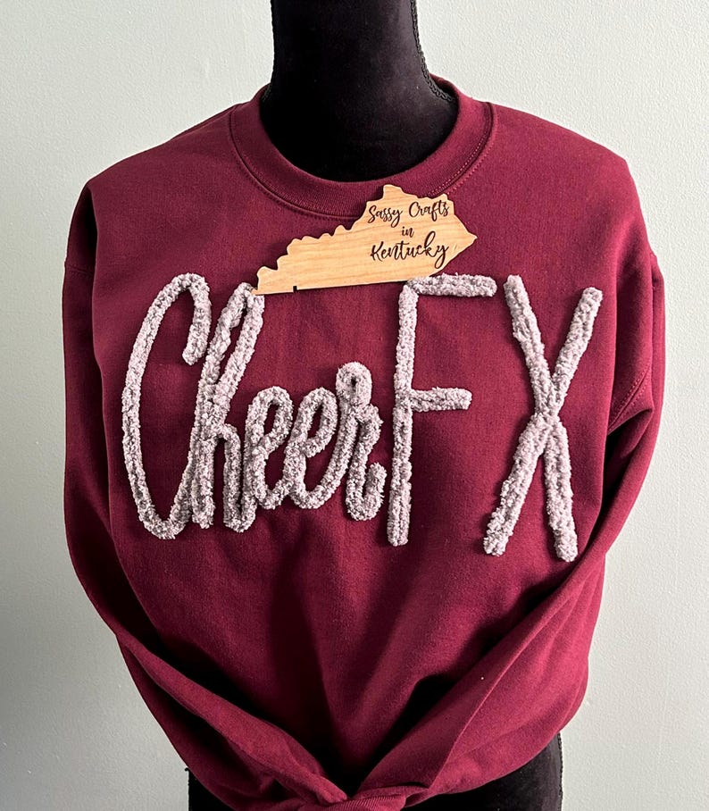May include: A maroon crewneck sweatshirt featuring the text CheerFX in raised, textured, light grey chenille-style lettering. A wooden cutout shaped like the state of Kentucky with the text Sassy Crafts in Kentucky is placed at the neckline.