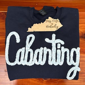 May include: A navy blue crewneck sweatshirt featuring the word Cabanting in large, light blue, textured chenille letters across the front. A wooden cutout shaped like the state of Kentucky with the text Sassy Crafts in Kentucky is placed above.