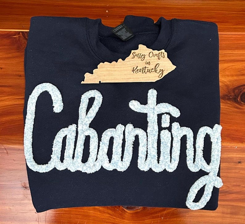 May include: A navy blue crewneck sweatshirt featuring the word Cabanting in large, light blue, textured chenille letters across the front. A wooden cutout shaped like the state of Kentucky with the text Sassy Crafts in Kentucky is placed above.