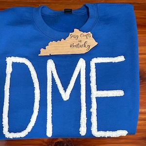 May include: A royal blue crewneck sweatshirt featuring the letters DME in large, white, textured chenille-style appliqu&eacute;. A wooden tag shaped like the state of Kentucky is placed on the chest, with the text Sussy Crafts in Kentucky engraved.