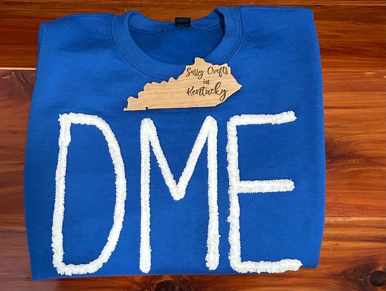 May include: A royal blue crewneck sweatshirt featuring the letters DME in large, white, textured chenille-style appliqu&eacute;. A wooden tag shaped like the state of Kentucky is placed on the chest, with the text Sussy Crafts in Kentucky engraved.